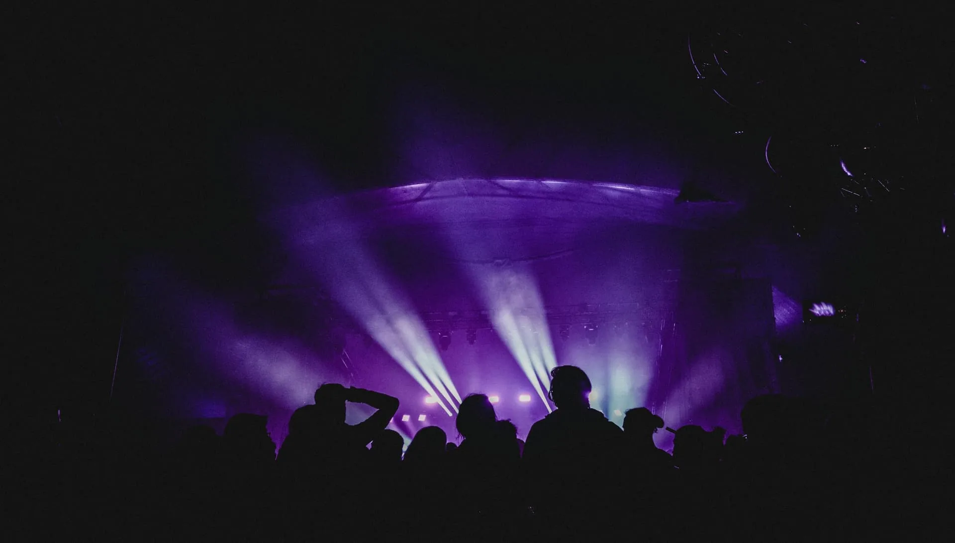Silhouetted crowd facing a stage with light beams cutting through purple haze